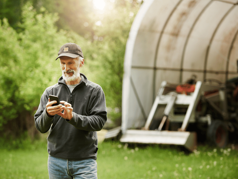 Farmer standing outdoors next to a greenhouse, holding a device, inspecting crops, with greenery and equipment in the background.